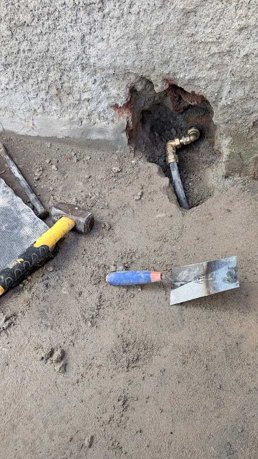 Tubazione riparata in una piccola apertura alla base della parete, con mazzuolo e cazzuola accanto